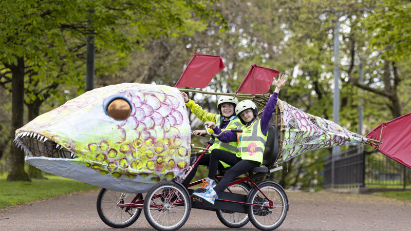 Two children ride a bike that is enclosed by a giant Paper mâché fish.