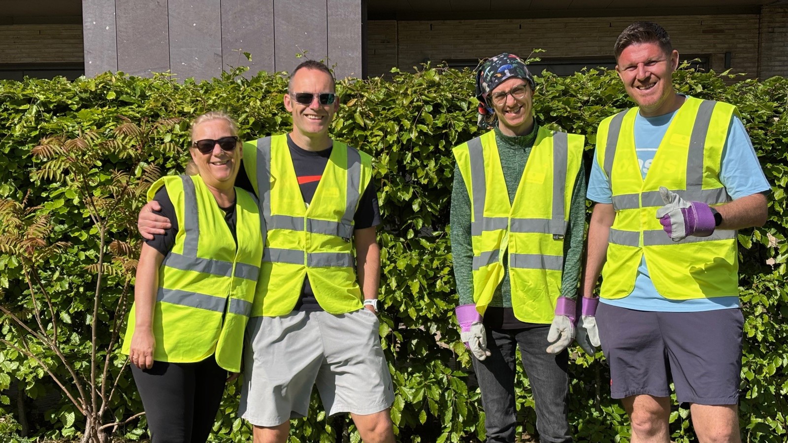 A group of four people wearing hi-vis vests and gardening gloves standing together in a sunny garden in front of a hospice building