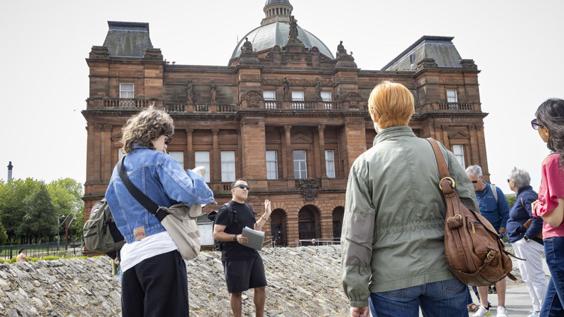 A tour guide wearing sunglasses with a tour group in front of the People's Palace entrance, they are wearing black, holding a clipboard and pointing to the museum.