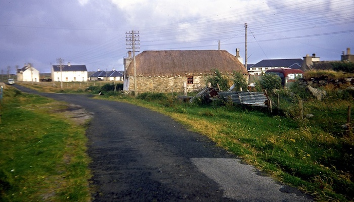 An old cottage with a thatched roof is in the foreground with a few white cottages in the background. A country road is shown and the image is from South Uist.