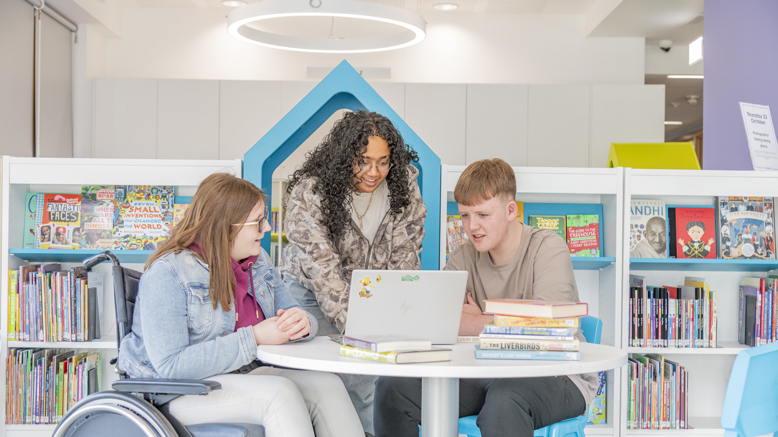 Three people are gathered round a table looking at a laptop, one is sitting in a wheelchair, one is sitting on a chair and one is standing. They are in a library