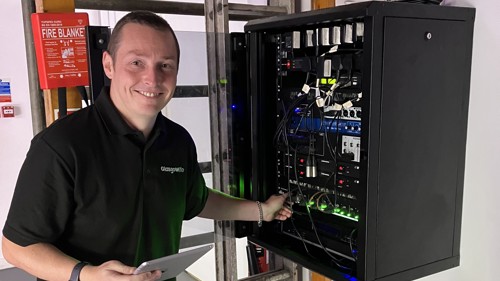 A person in a black polo shirt is standing next to a control panel for a PA system in a community centre