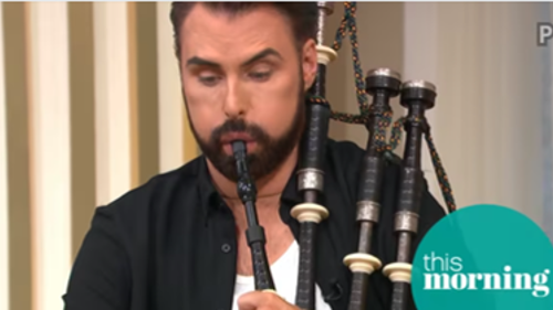 A person with dark hair is blowing into a set of bagpipes on a sofa in a tv studio