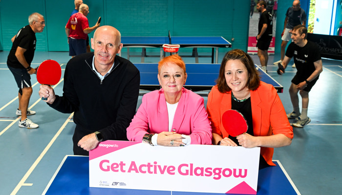 A man and woman holding table tennis bats stand either side of a woman wearing a pink jacket leaning on a sign which reads 'Get Active Glasgow'