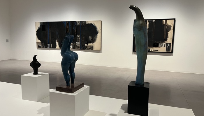  A bright white walled gallery space displays three ceramic sculptures on white pedestals. The central sculpture resembles a stylised blue human torso with soft curves, the head and arms are missing. The ceramic sculptures on either side resemble abstract bird forms with the one on the left tall and slender and the one on the right small and dark with a smooth, rounded form. Behind them hang two large abstract paintings featuring dark, textured surfaces with bare canvas and photographic fragments