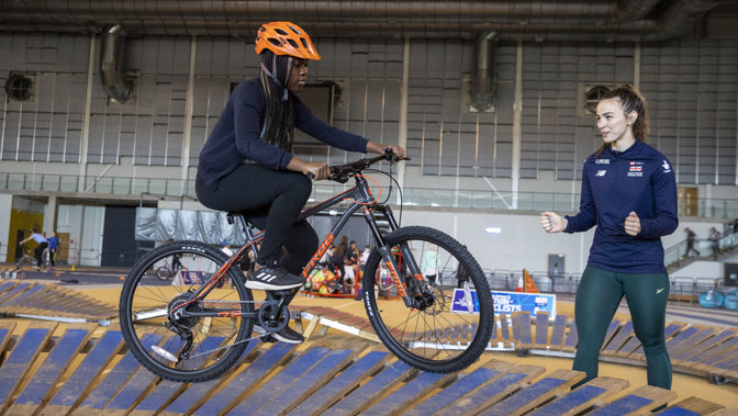A person wearing a helmet on a bike cycling around a blue track, a coach is to the right of the picture