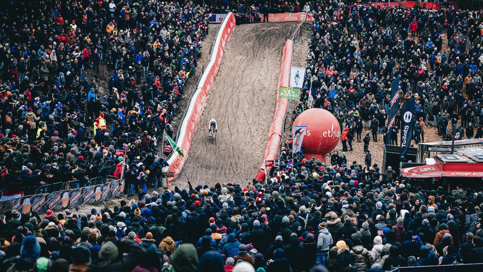 A large crowd in watching an outdoor cycling event in a park. A cyclist is racing on a sandy, hilly track