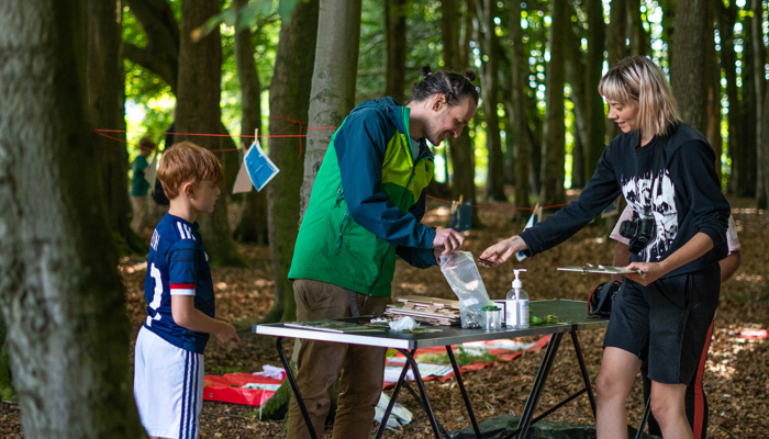 Three people stand in a wood they are smiling and looking at drawings which are displayed on a table.