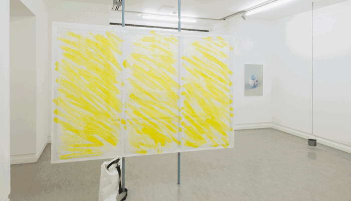 A three-panel fine mesh covered screen painted with three blocks of bold yellow brushstrokes stands in a white gallery, with a small hanging object and wall artwork in the background.