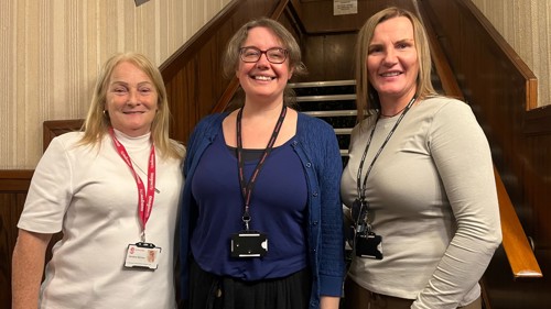 A group of three people standing and smiling at the bottom of a wooden staircase in an historic community building