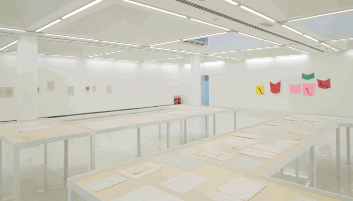 A bright, minimalist gallery with long white display tables covered in papers, surrounded by sparse wall works and a row of small, colourful fabric artworks on one wall.