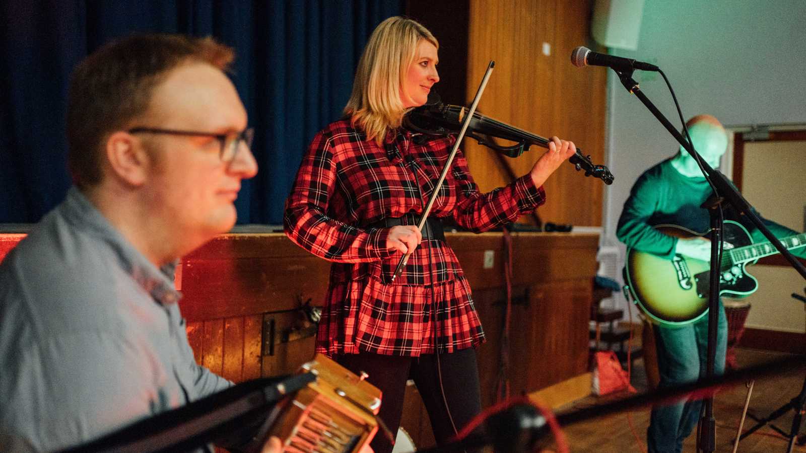 Three musicians play an accordion, violin and guitar in a community setting