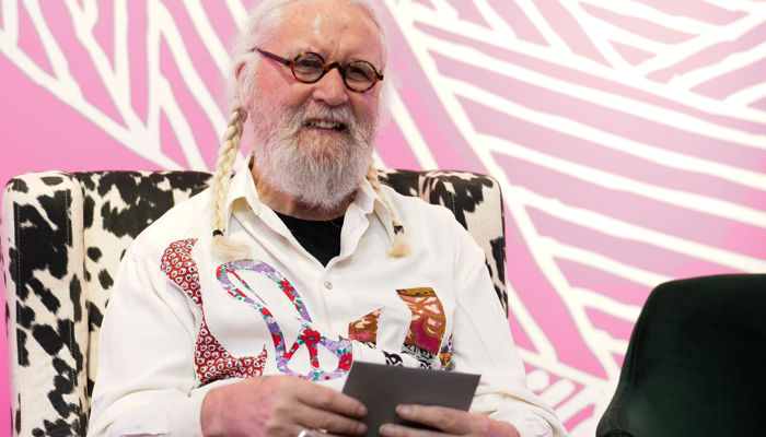 Billy Connolly sitting in a chair with a pink background looking and smiling to the front