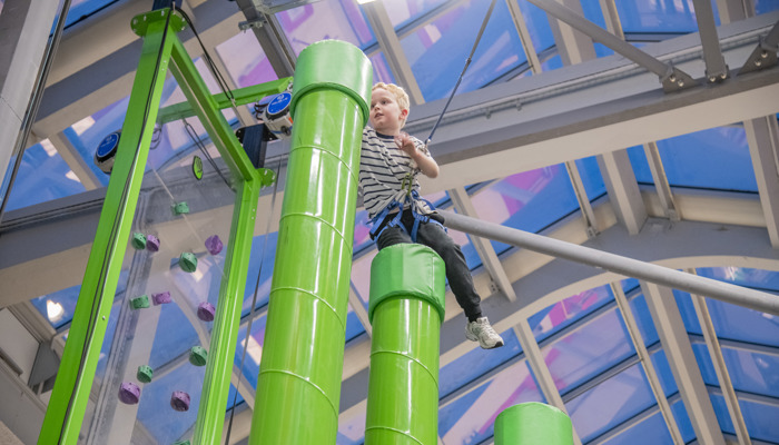 A young boy with a stripy top and harness climbing up green cylinders with each on getting higher.