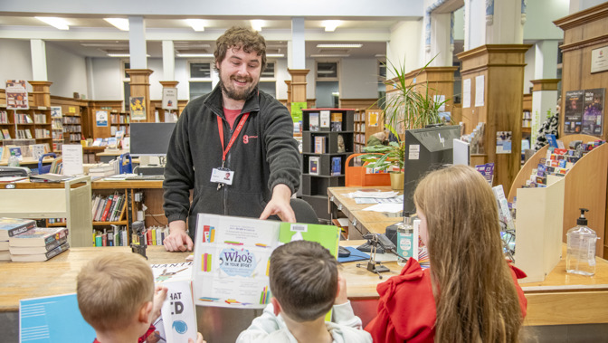 Glasgow Libraries staff member behind a wooden counter hands a children’s book to three children standing on the opposite side. The library interior includes bookshelves, display stands, and computer equipment, with bright overhead lighting. A sign on the counter display reads “Who’s in your book?” and other books are stacked nearby.