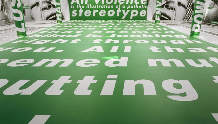 A large-scale installation in a bright, modern gallery space dominated by vivid green surfaces. The floor, central wall, and several cylindrical columns are all covered in bold white sans-serif text set against a green background. The perspective is low and wide, with oversized words stretching across the floor toward the viewer, creating a strong sense of depth and immersion. On the far wall, large white lettering reads: “All violence is the illustration of a pathetic stereotype”.