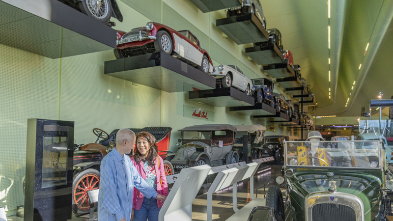 Visitors explore the Riverside museum in Glasgow, standing at an interactive display in a large gallery filled with classic automobiles, including early 20th‑century vehicles on the floor and additional cars mounted on tiered wall shelves above.