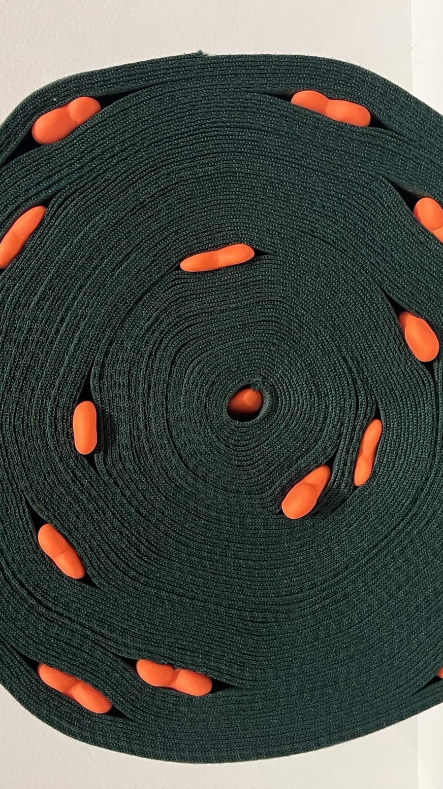 A tightly coiled roll of dark green material forms a neat spiral against a light wall. Small bright orange bean shapes are embedded at regular intervals along the coil, creating a strong colour contrast. The lighting highlights the texture of the fabric and the precise, geometric pattern of the spiral.