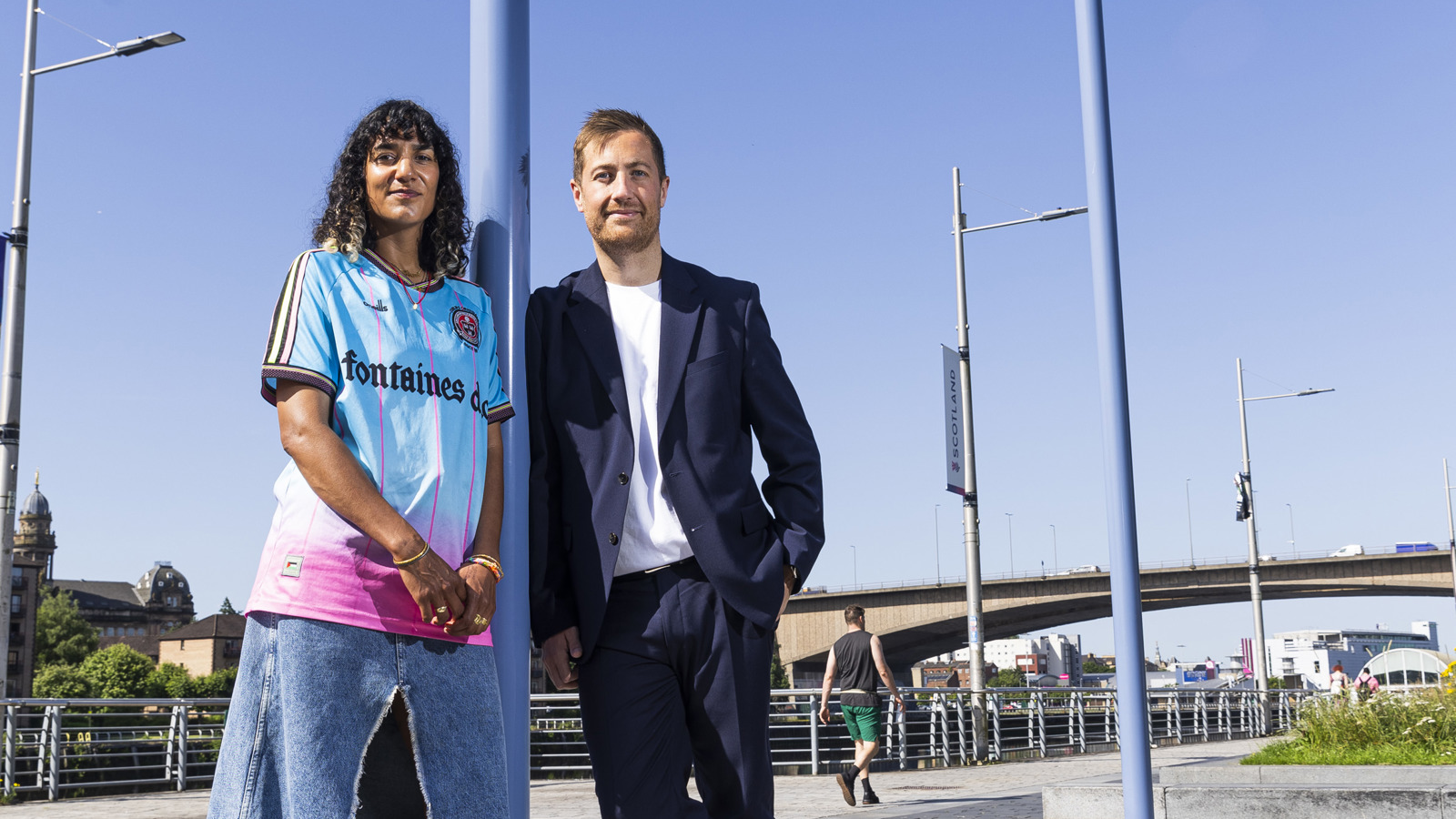 A woman and man lean against a pole blue, with a blue sky and bridge in the background