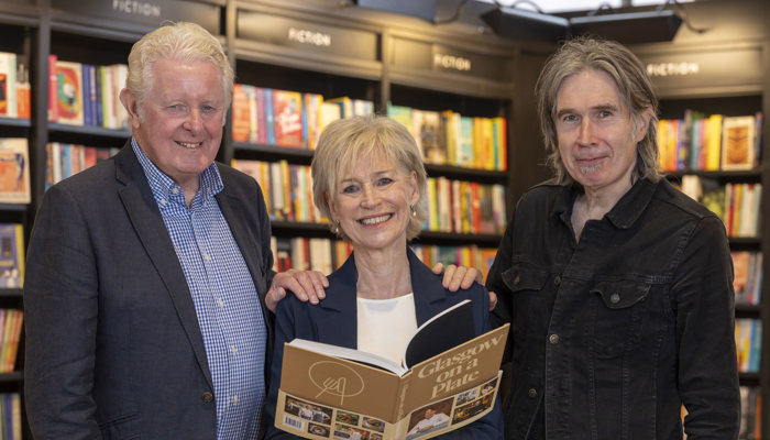 Two men and a woman stand in a classically furnished library. The woman is standing in the middle of the three holding a book