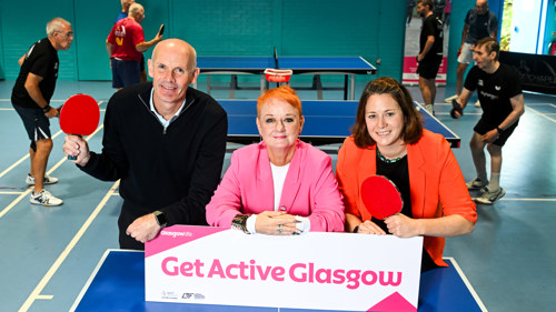 Three people are standing behind a table tennis table in a sports hall with people playing the game behind them. Two of them are holding table tennis bats. They have a sign in front of them that reads Get Active Glasgow
