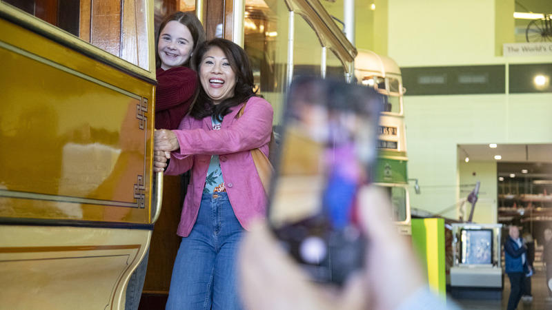 Two people lean out of the open doorway of a vintage yellow and wooden tram inside Riverside museum in Glasgow. Another tram is visible behind them, and a blurred smartphone held in the foreground suggests a photo being taken.