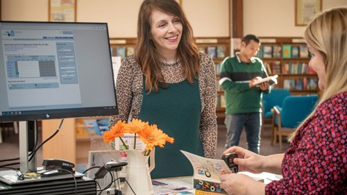 A person smiling and checking out a book at a library counter. The Librarian is stamping the book