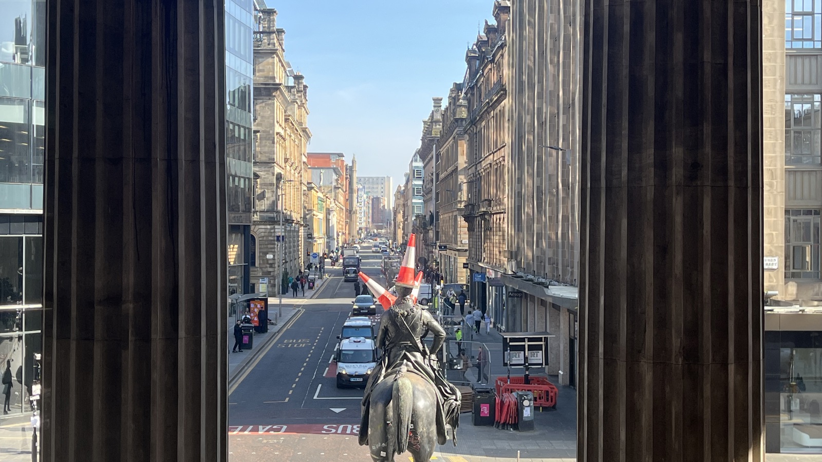A city street is viewed between two large stone columns, with an equestrian statue centered in the foreground. The rider on the statue has two orange traffic cones placed on their head, creating a humorous contrast with the otherwise grand scene. Historic and modern buildings line the busy street behind it, where pedestrians and cars move through the urban settin