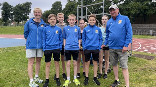 Two adult coaches from Shettleston Harriers with a group of young athletes, who are a mix of genders, at Crownpoint athletics track in Glasgow.