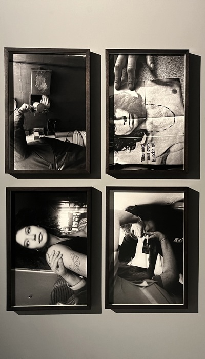 A gallery wall displays four framed black‑and‑white photographs arranged in a two‑by‑two grid. The top-right image shows a person transferring liquid from their cup into a cup held through a gap in a prison door. The top-left image focuses has a young woman being led from one room to another. The bottom-right image shows a handing holding down a poster of Jim Morrison of The Doors. The bottom-left image shows a women taking a breathalyser test. Below a grid of four labels has information about the works.