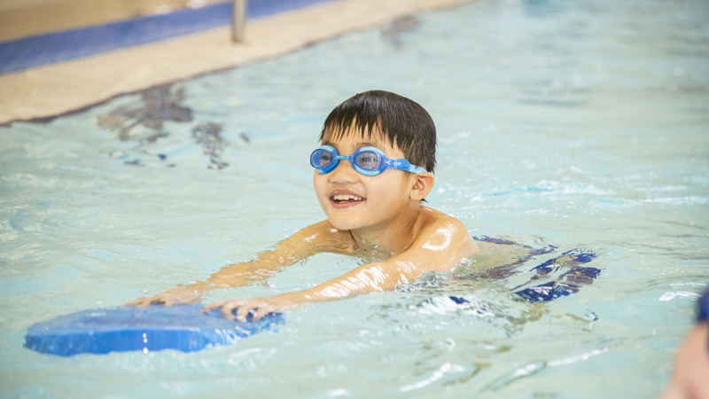 A child wearing swim goggles practices kicking in a swimming pool while holding a blue kickboard, gliding forward through the water near the pool’s edge.