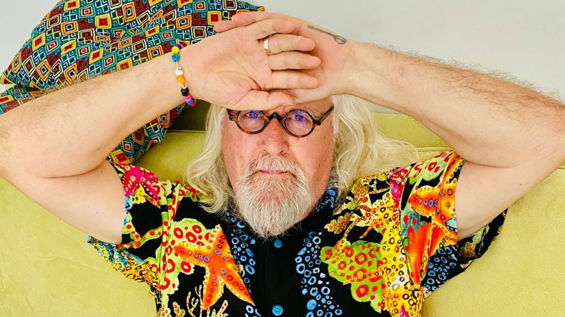 A close up of Sir Billy Connolly with his long, light-coloured hair and a beard reclining on a light-coloured sofa, resting both hands across his forehead. He wears round glasses, a brightly patterned shirt with bold colours, a bracelet, and a ring. A cushion with a geometric pattern is visible behind his head against a plain background.