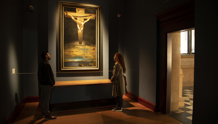 Photograph of two people standing in a dimly lit room. They are looking at a painting which is spot lit. The painting shows a biblical scene, the crucifixion of Jesus from the artists perspective.