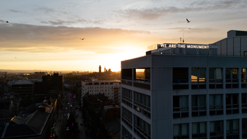 A large illuminated sign that reads We Are The Monument on top of a tall concrete building in a city centre at dusk