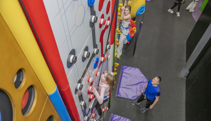 A young girl who is wearing a pink top scaling a multi-coloured climbing wall.