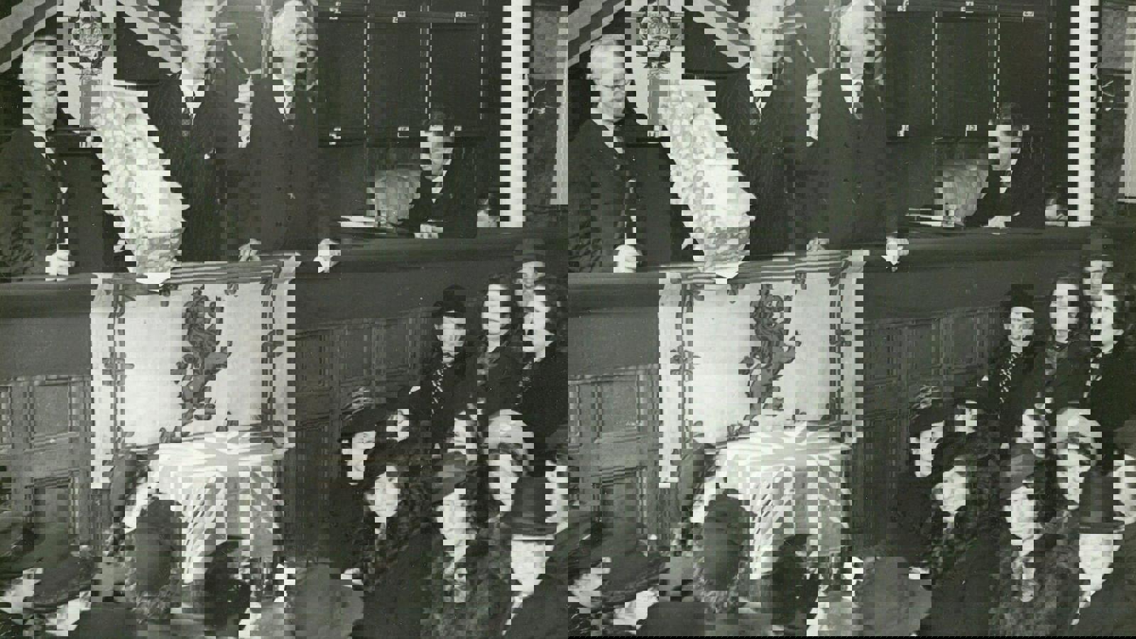 Three people standing talking to a crowd lower below, holding a baby in a small crib with a Lion Rampant of Scotland flag and a Scotland flag draped below.