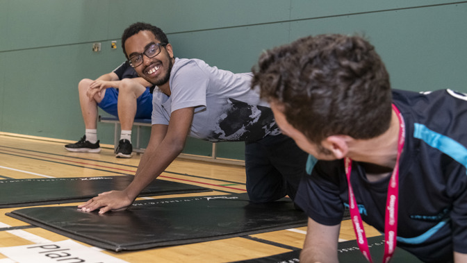 Two men are smiling and exercising on mats on the floor of a sports hall.