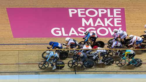 Cyclists on an indoor track ride past a pink and white sign which reads 'People Make Glasgow'