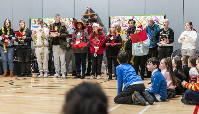 A group of people of various ages sing as choir in a school hall. 