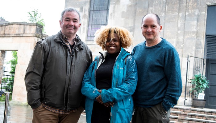 Three figures stand together in front of a brick building 