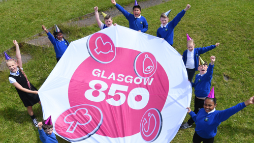 A group of young people in blue school uniform and colourful party hats are holding up a large round banner between them that reads Glasgow 850. They are cheering and holding their other arms up in the air