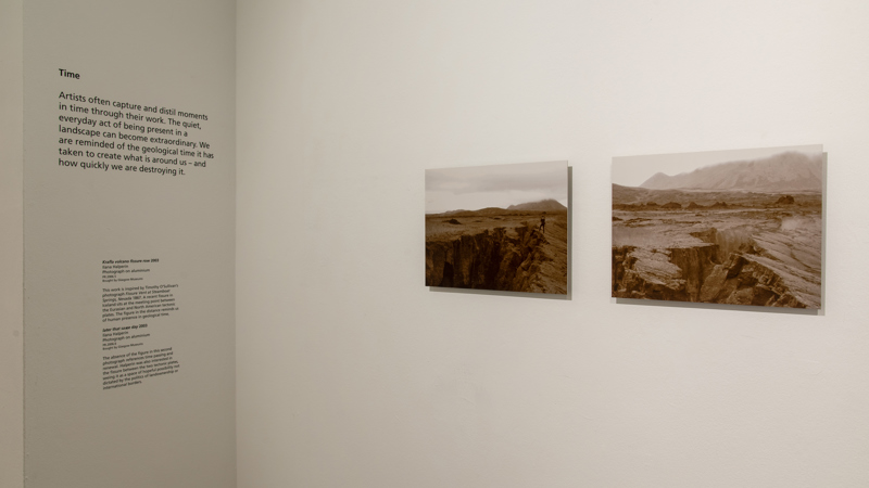 A corner of gallery with white walls. One wall has two exhibition texts and the other two sepia photographs of rugged landscapes mounted on aluminium and hung on the wall