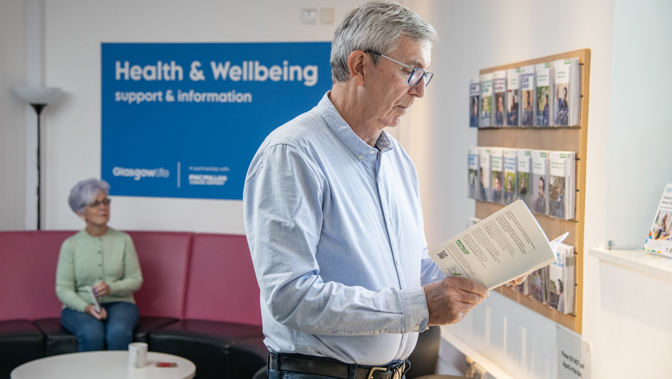 A man with glasses standing and reading a book, another person is sitting down behind him.