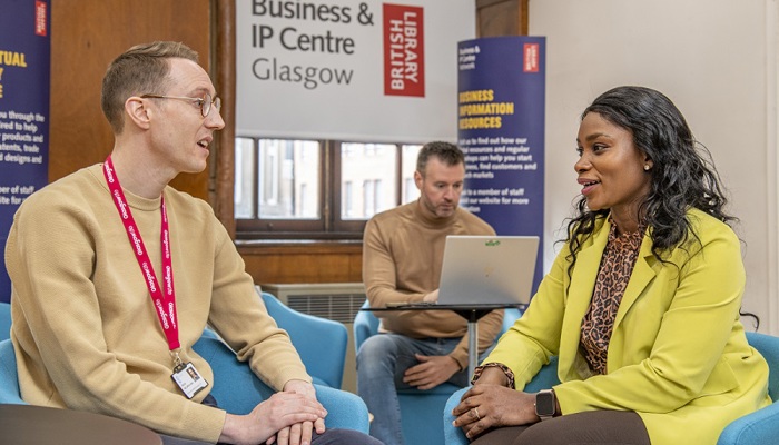 Two people sitting in armchairs having a conversation in a library. There is a person working on a laptop behind them.