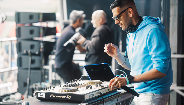 A DJ performs on stage using a mixing deck and laptop, holding a microphone, while sound equipment and event staff are visible in the background.