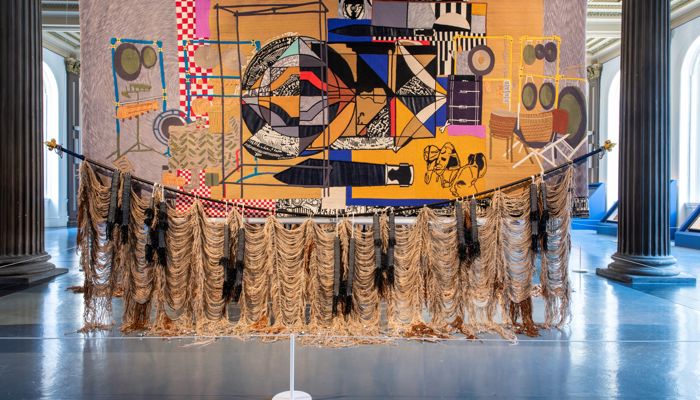 A large, richly detailed textile artwork with geometric and figurative patterns hangs in a grand columned hall, with long strands of rope-like fringe draping along its lower edge.