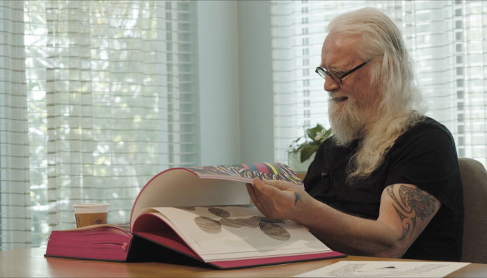 Billy Connolly sitting at a desk in a room with slatted blinds in the window, he is holding a large book and is turning the pages