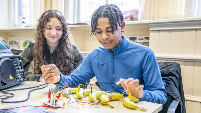 Two people sit at a table in a classroom, working on an electronics project using a small circuit board connected with wires to several bananas. One person holds a smartphone while the other interacts with the wired setup. Tools, cables, and equipment are spread across the table, with large windows and classroom furnishings visible in the background.