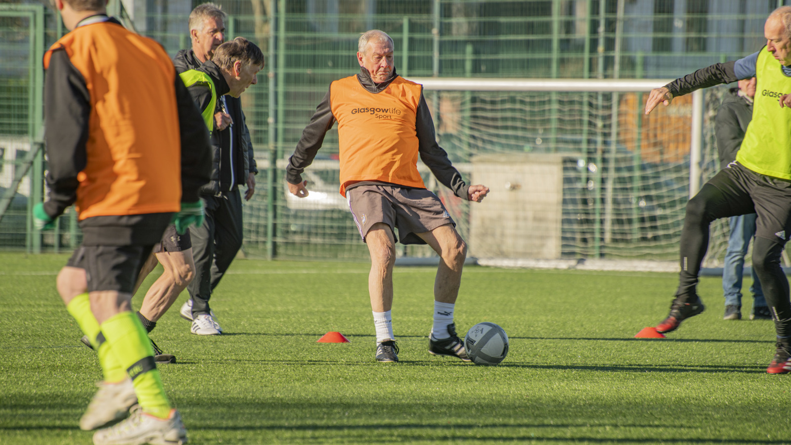 People wearing brightly coloured bibs during a game of Walking Football outside on an artificial grass pitch.