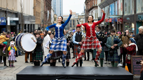 A photo of two highland dancers in traditional dress jumping in the air, with a piping band playing behind them. Photo credit: Alan Harvey, SNS Group 